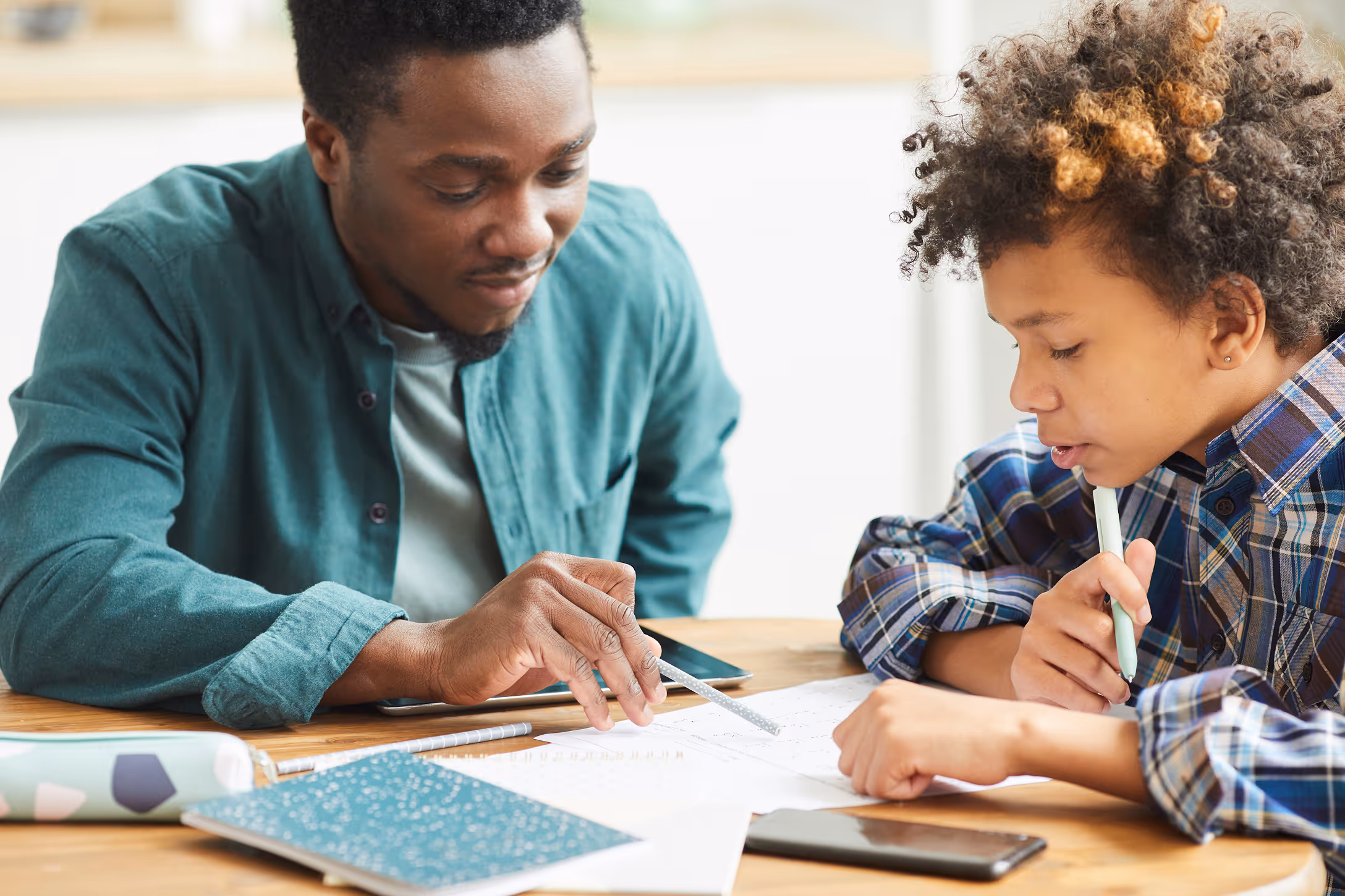 An adult and a child sitting at a table, working together on homework with notebooks and a smartphone nearby.