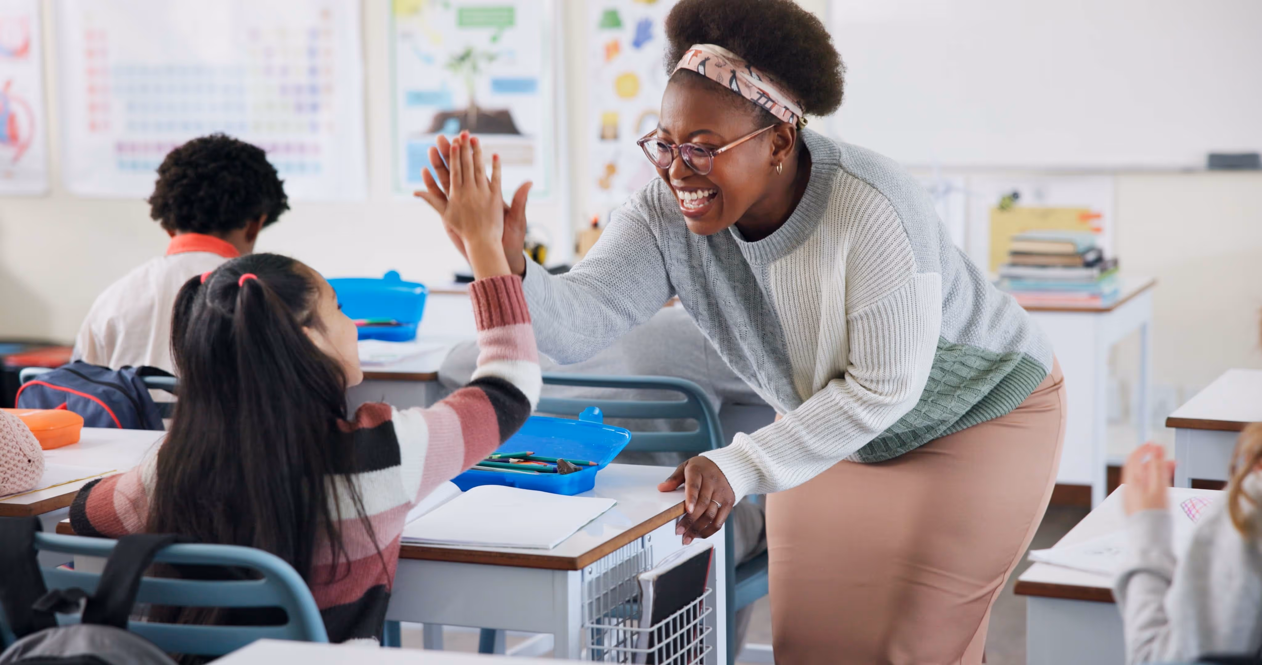 Smiling teacher giving a high-five to a seated girl in a classroom with other students.