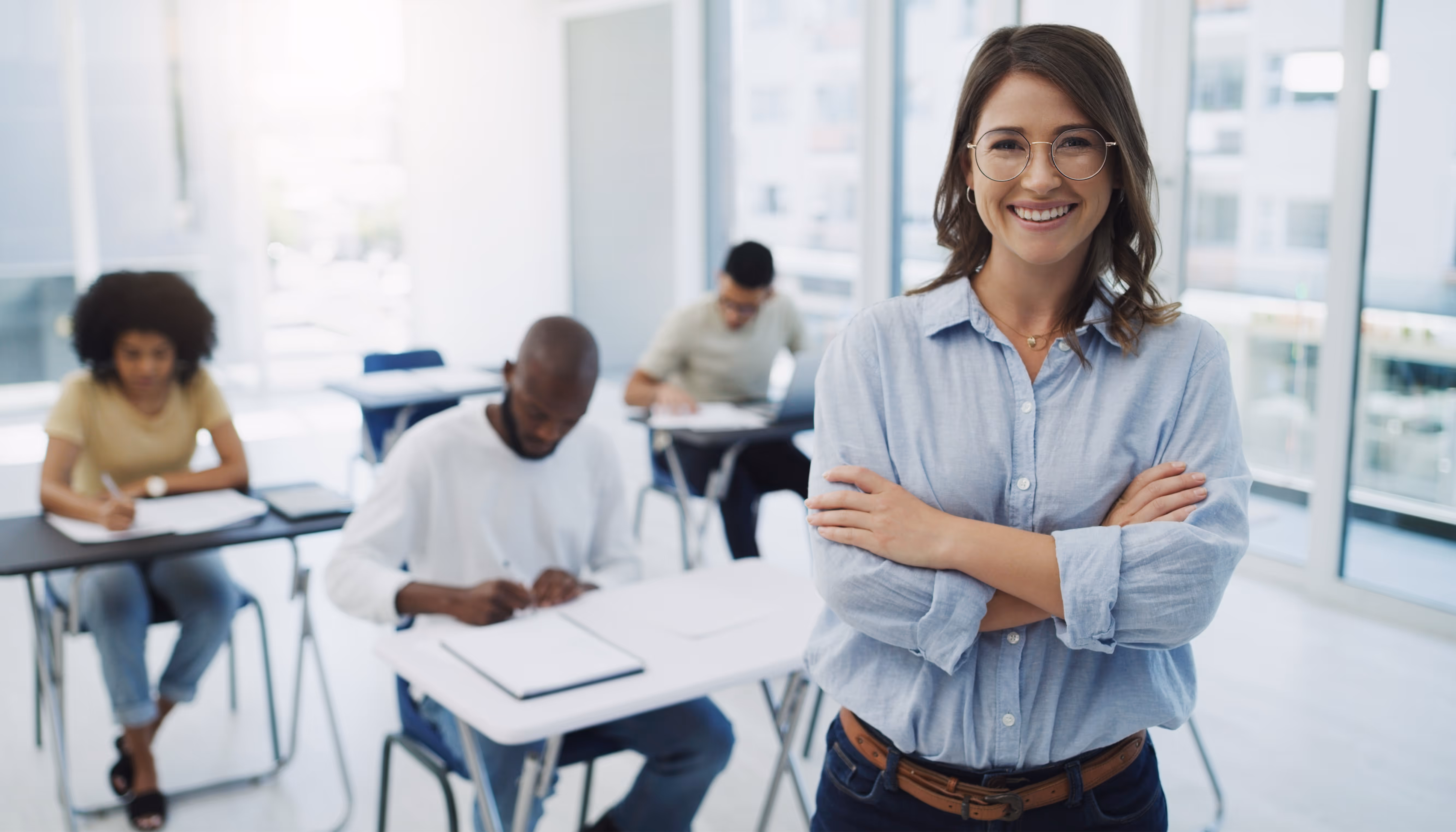 Smiling female teacher with glasses stands with arms crossed in front of seated students writing in classroom.