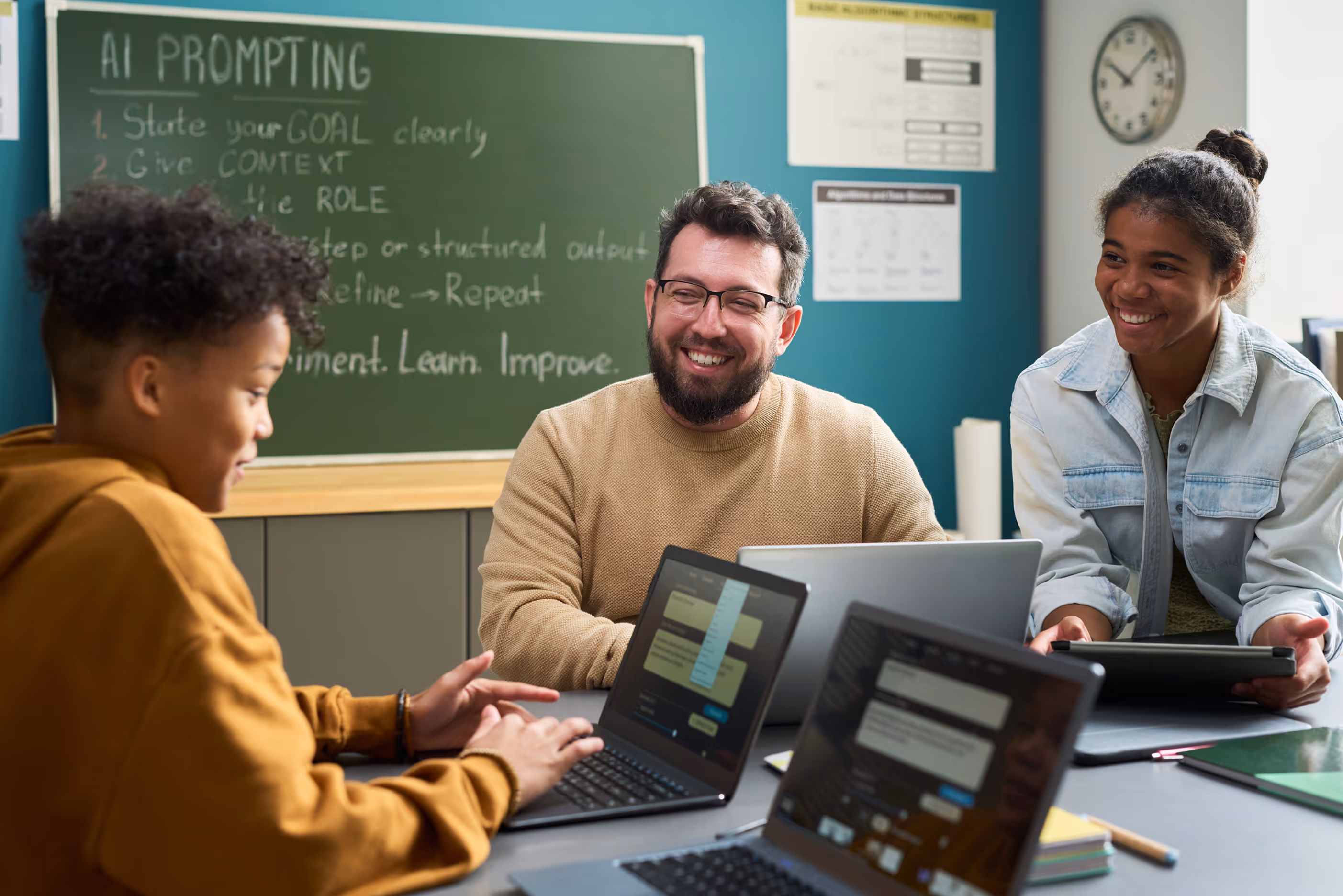 Three people sit at a table with laptops in a classroom with AI prompting instructions on a chalkboard.