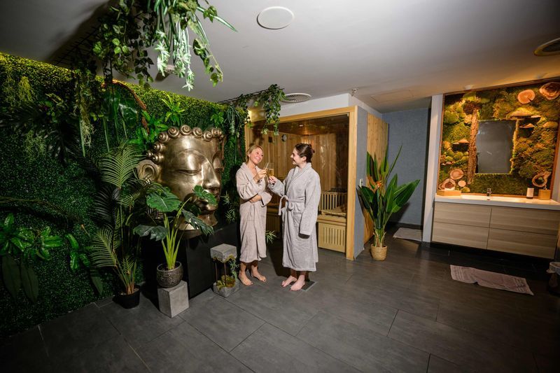 Deux femmes en peignoir trinquent avec des coupes de bulles devant un sauna finlandais, dans l'espace Spa Nature de DAO Liège