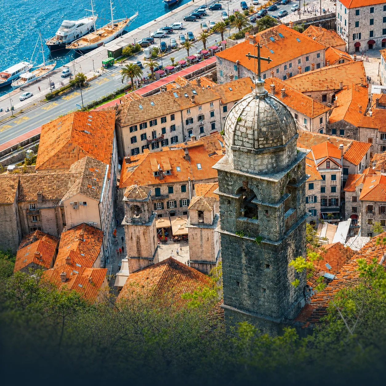 Aerial view of a historic old town with orange-tiled roofs, narrow streets, a prominent stone church tower with a cross in the foreground, and a waterfront with docks and boats in the background.
