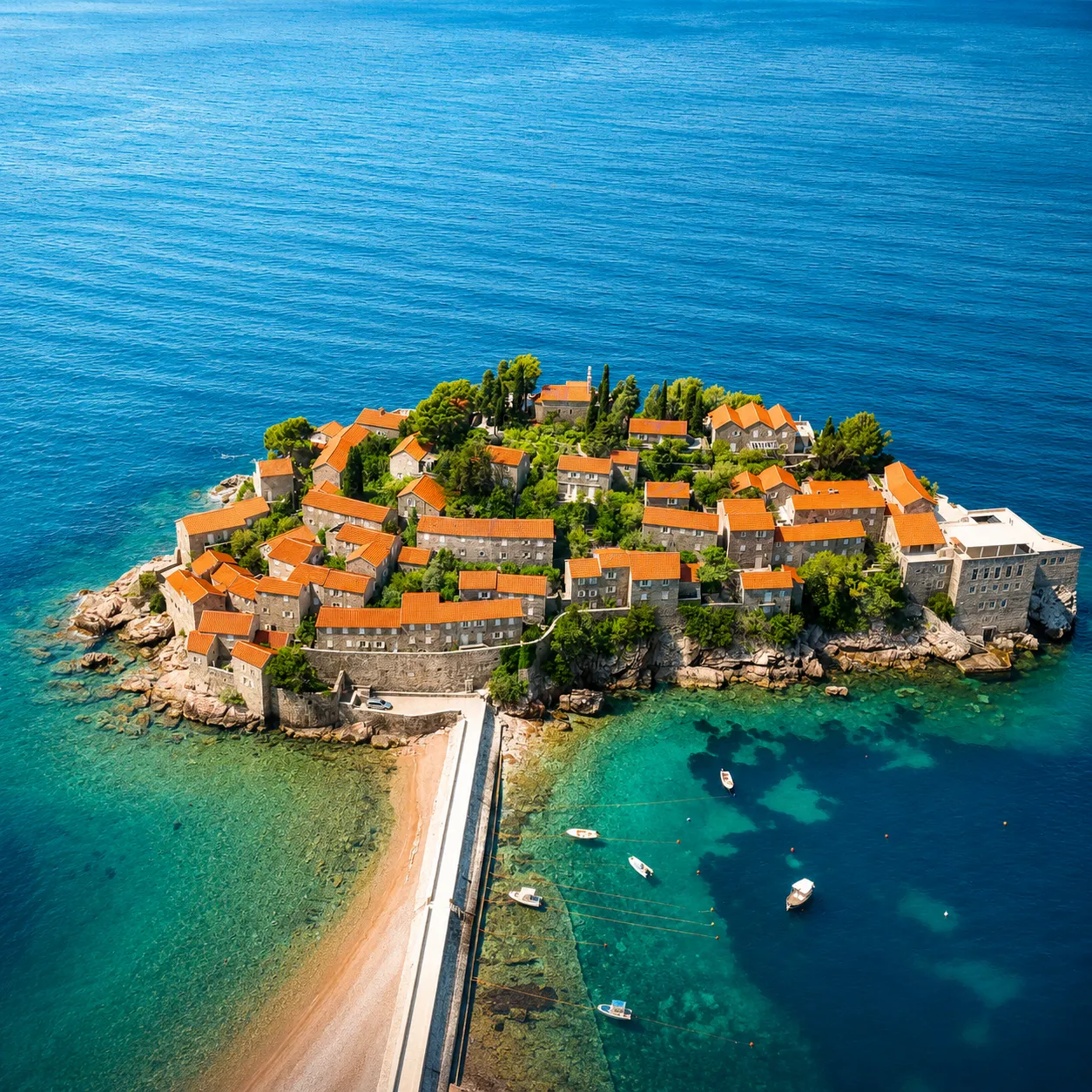 Aerial view of a small island featuring clustered stone buildings with orange roofs, surrounded by clear blue sea, connected to the mainland by a narrow causeway, with several small boats moored near the shore.