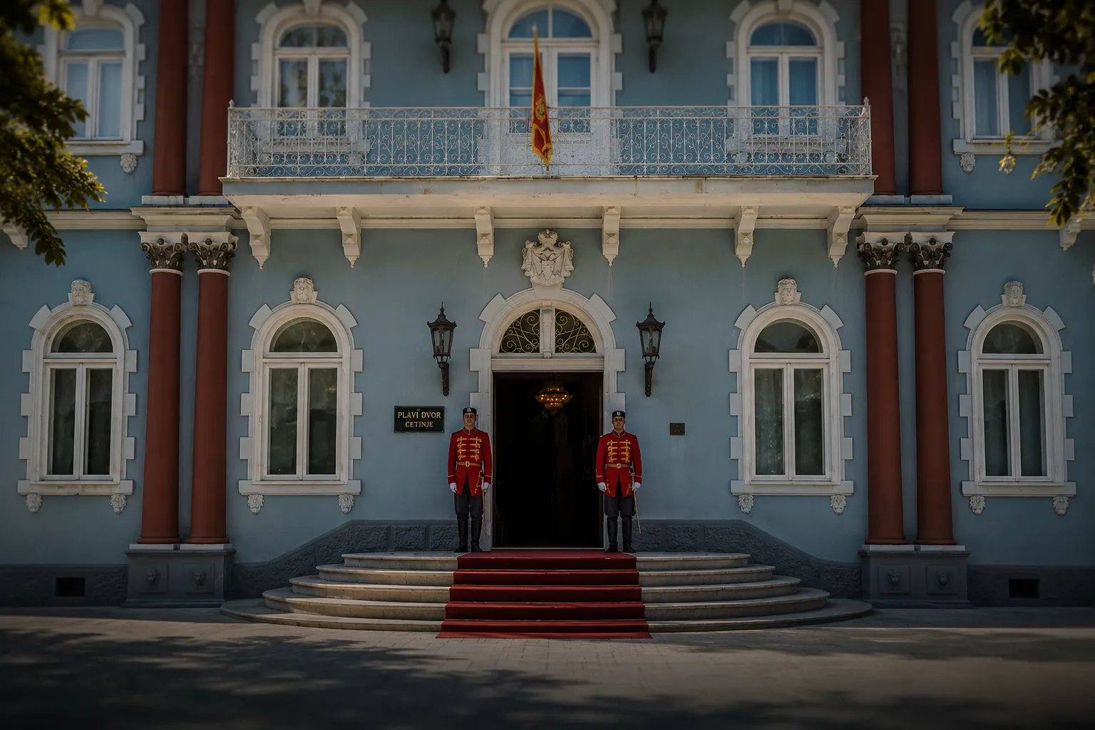 Front view of an elegant blue building with white-trimmed arched windows and red columns, featuring a central arched entrance with two uniformed guards standing on either side of a red carpeted staircase, and a balcony with a flag above.