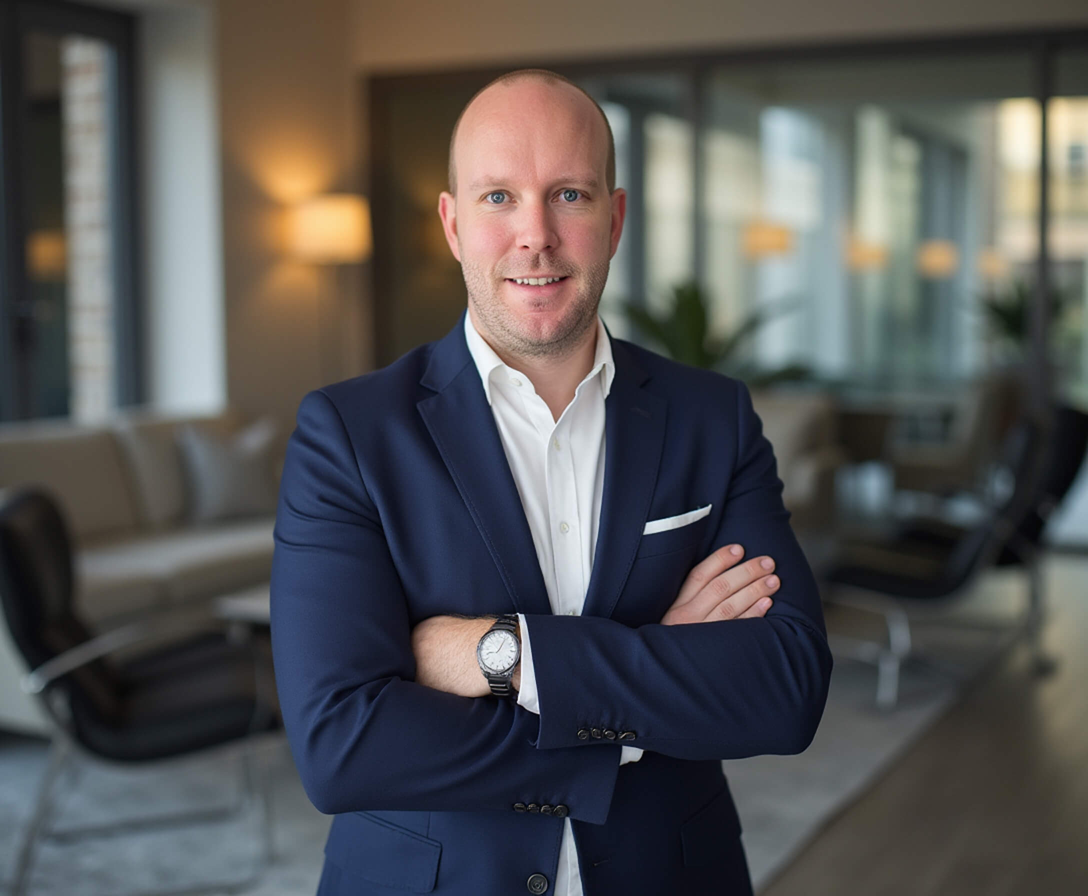 Markus Berger in einem dunkelblauen Anzug mit weißem Hemd steht mit verschränkten Armen in einem modernen Büro mit Sitzmöbeln und warmem Licht.