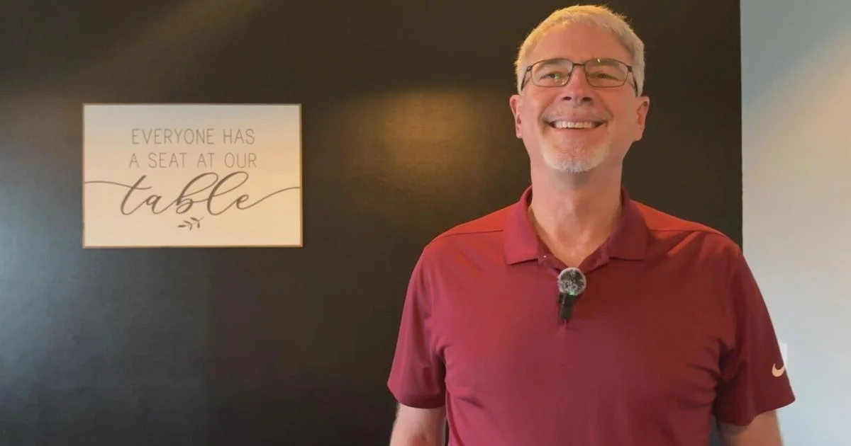 Smiling Dean with glasses and gray hair wearing a red polo shirt standing in front of a black wall with a sign that says 'Everyone has a seat at our table'.
