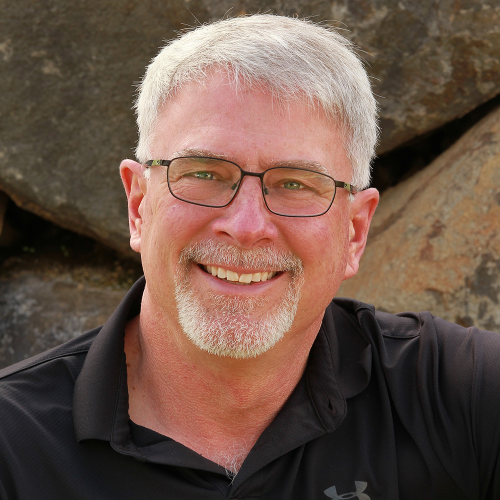 Smiling Dean with gray hair, glasses, and a goatee wearing a black Under Armour polo shirt sitting outdoors in front of large rocks.