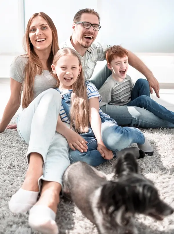 Happy family of four sitting on a carpet recently cleaned by Dean with Valley Professional Carpet with a small black and gray dog in the foreground.