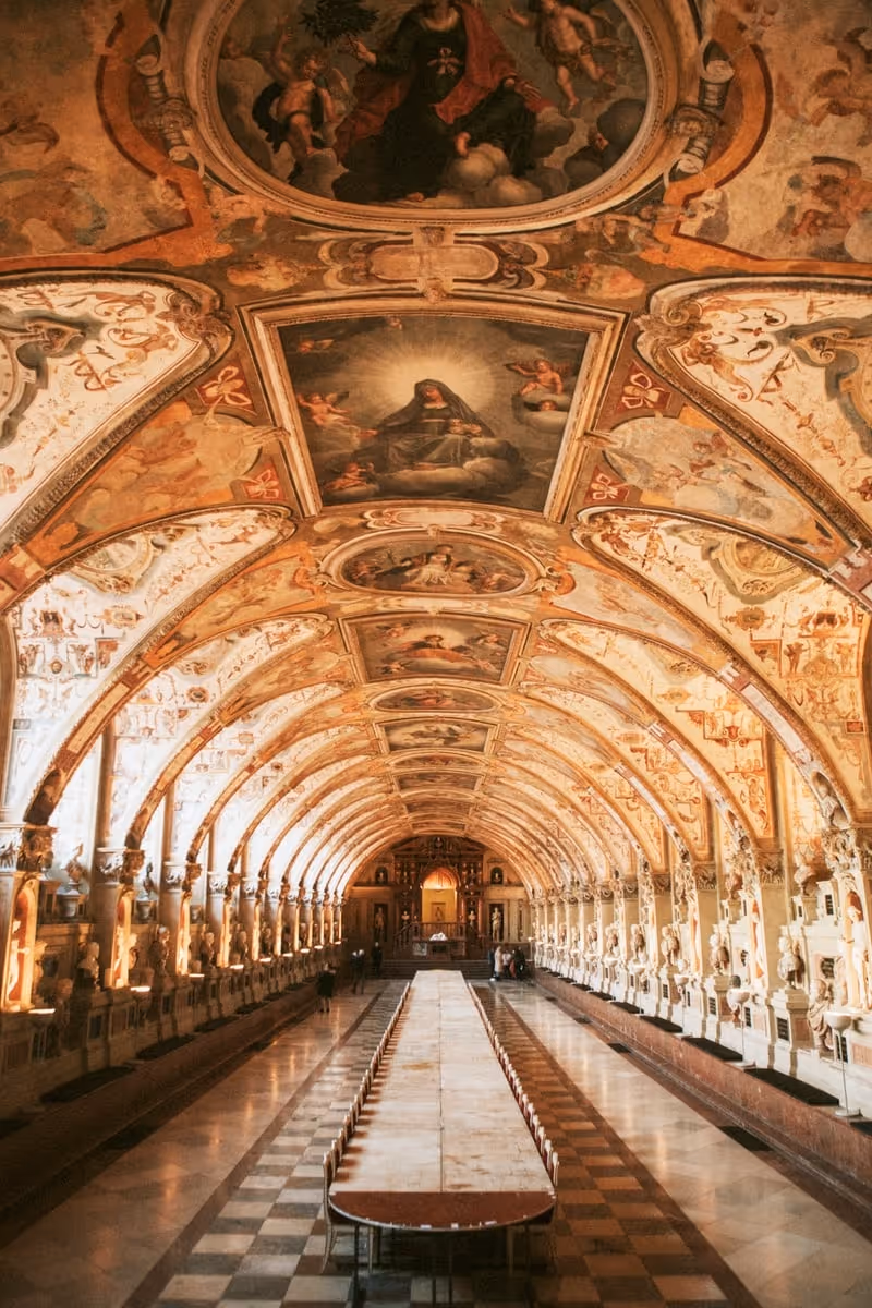 Long ornate hallway with vaulted ceiling decorated with religious frescoes and sculptures lining the sides, featuring a central long table and chairs.
