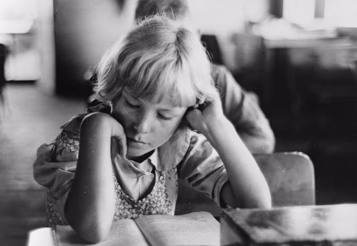 Black and white photo of a young child with short blond hair intently reading a book indoors.