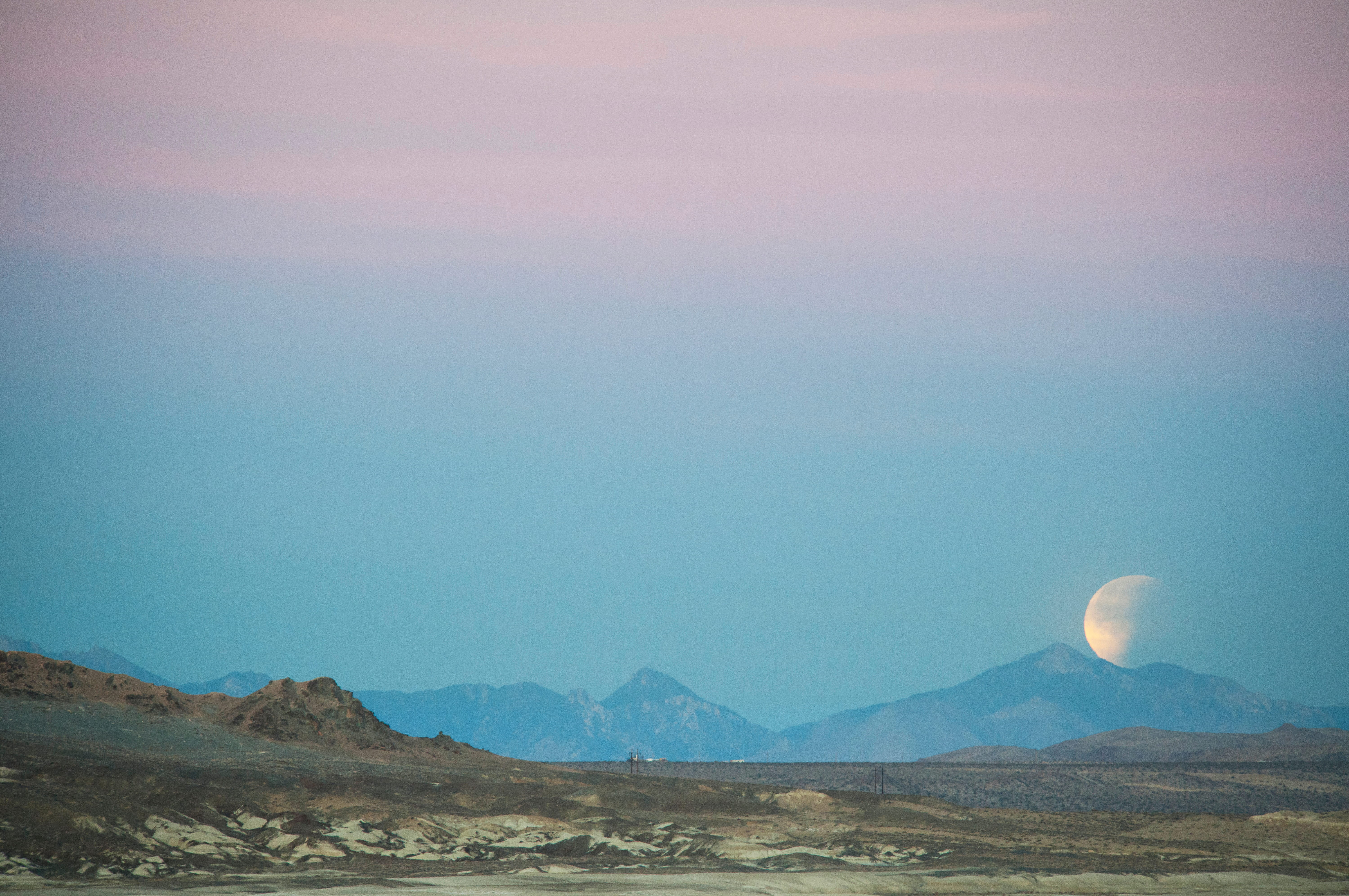 Half moon rising behind mountain range under a pastel sky transitioning from pink to blue.