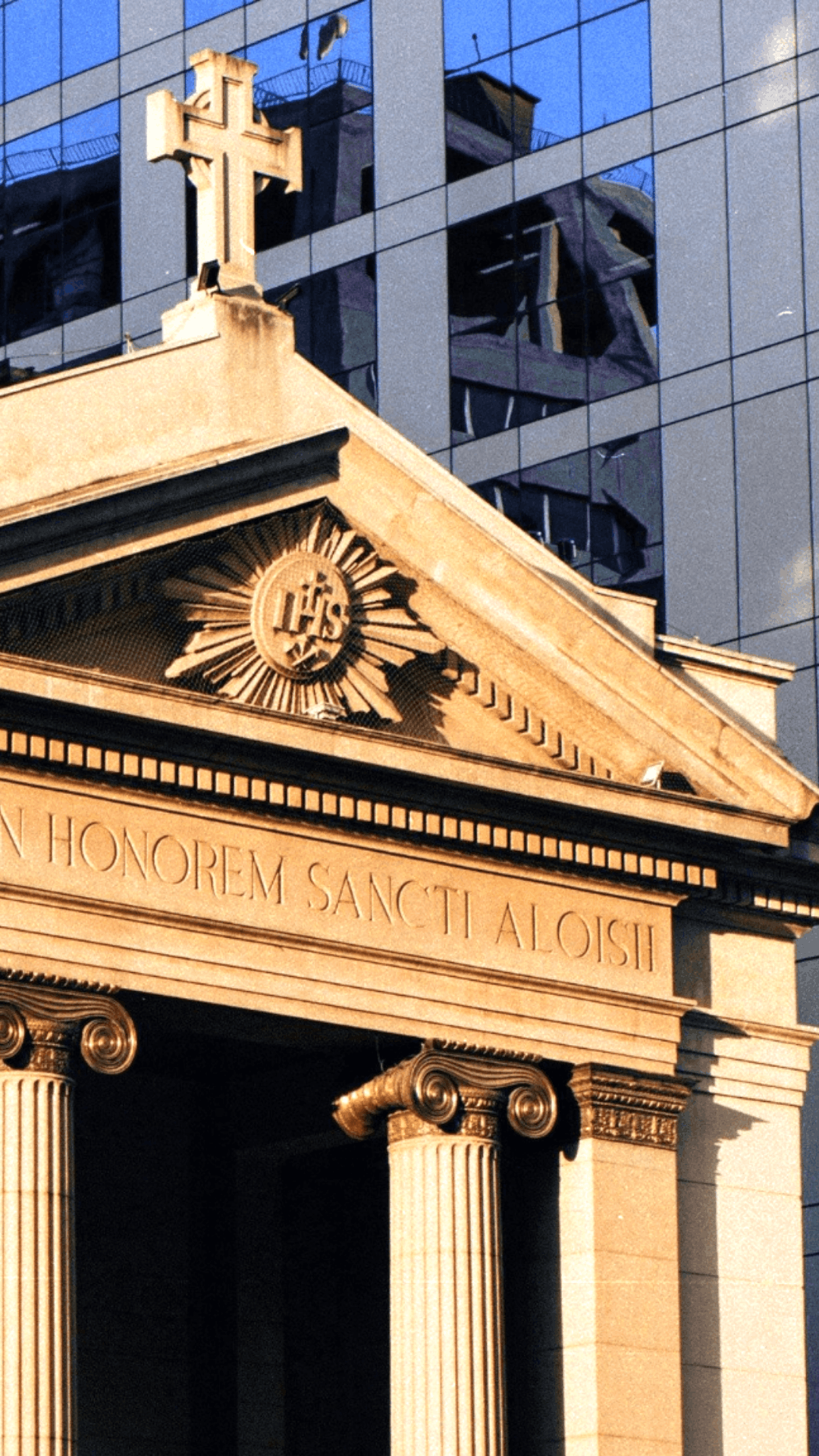 Classical building facade with decorative columns, a Latin inscription, and a cross on top against a modern glass skyscraper background.