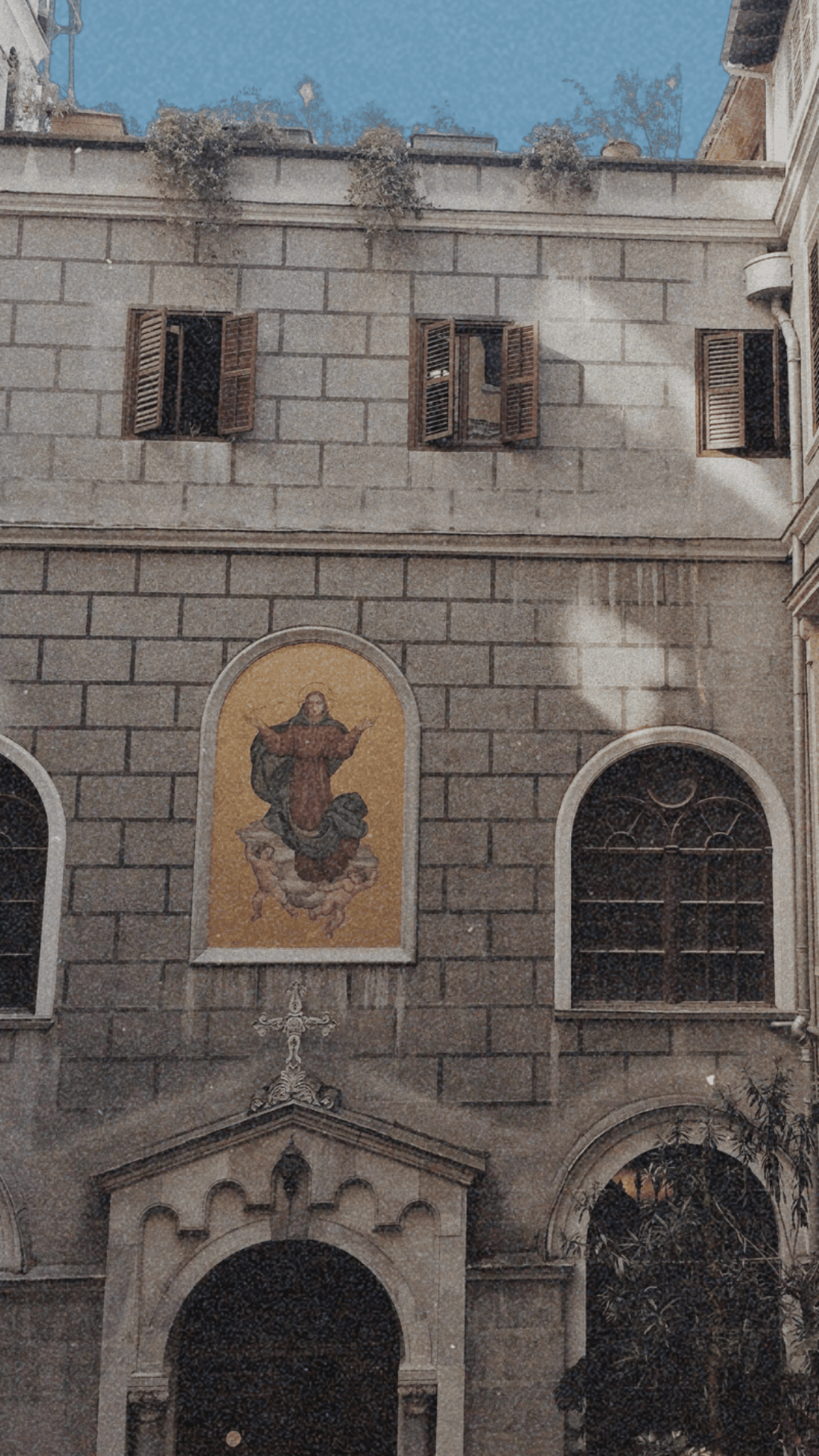Stone building facade with open wooden shutters, a religious mural of a saint above the entrance, and a decorative stone archway with a cross on top.