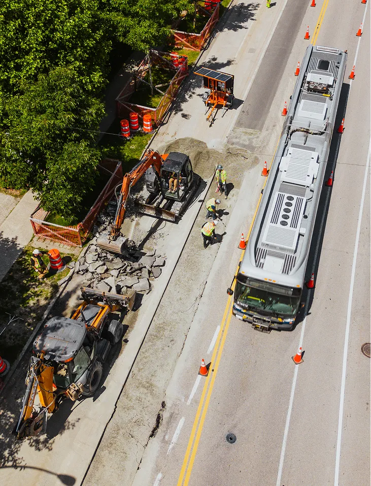 Road construction with workers, excavator, loader, and a bus beside traffic cones on a sunny day.