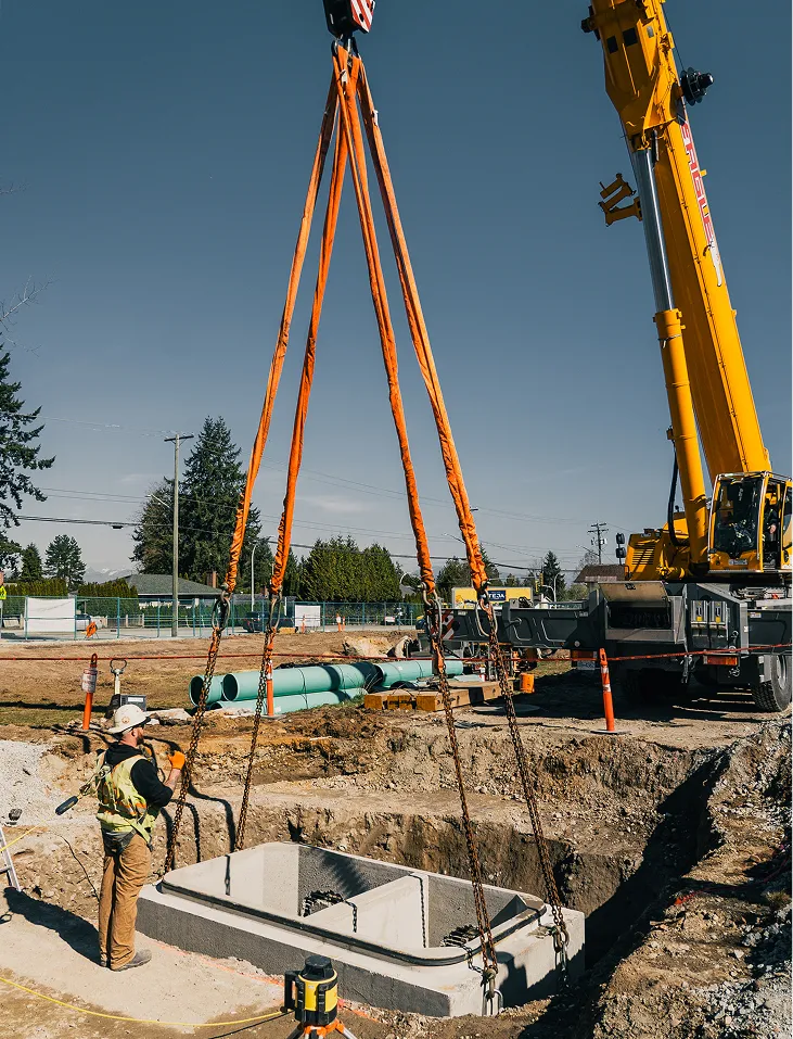 Construction worker guiding a concrete structure being lowered into a trench by a crane with orange slings.