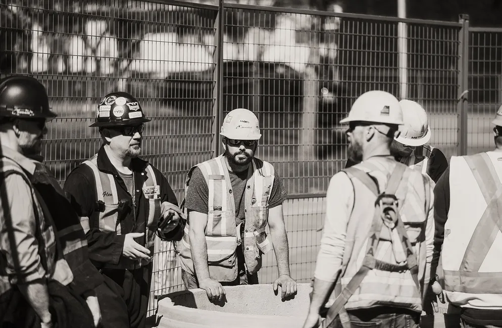 Group of construction workers wearing hard hats and safety vests at a fenced site.