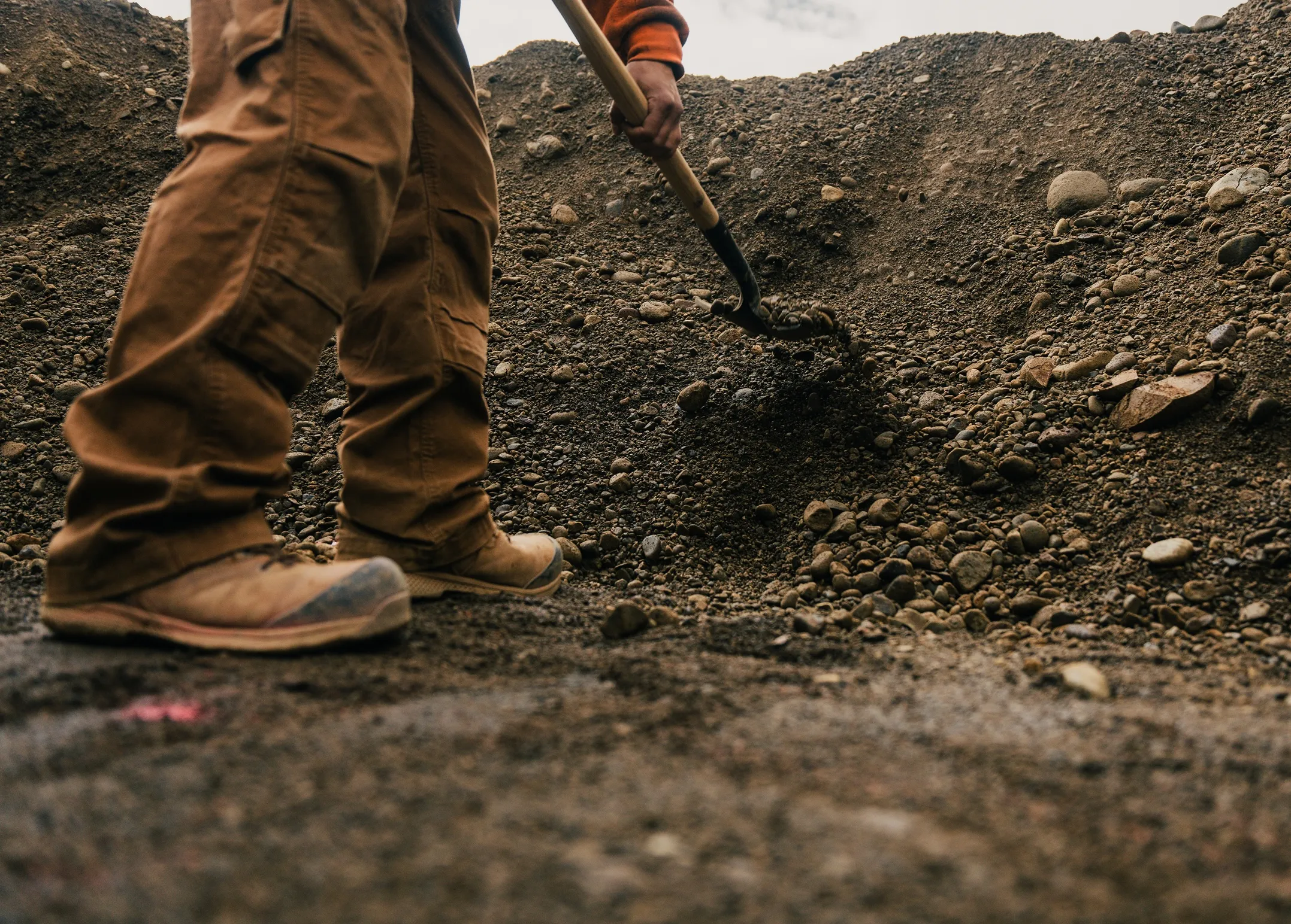 Worker in brown pants and boots shoveling gravel or dirt at a construction site.