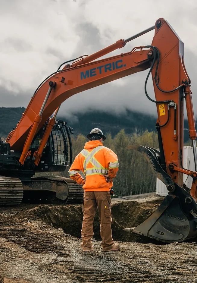 Construction worker in orange safety gear standing near an orange excavator labeled METRIC.