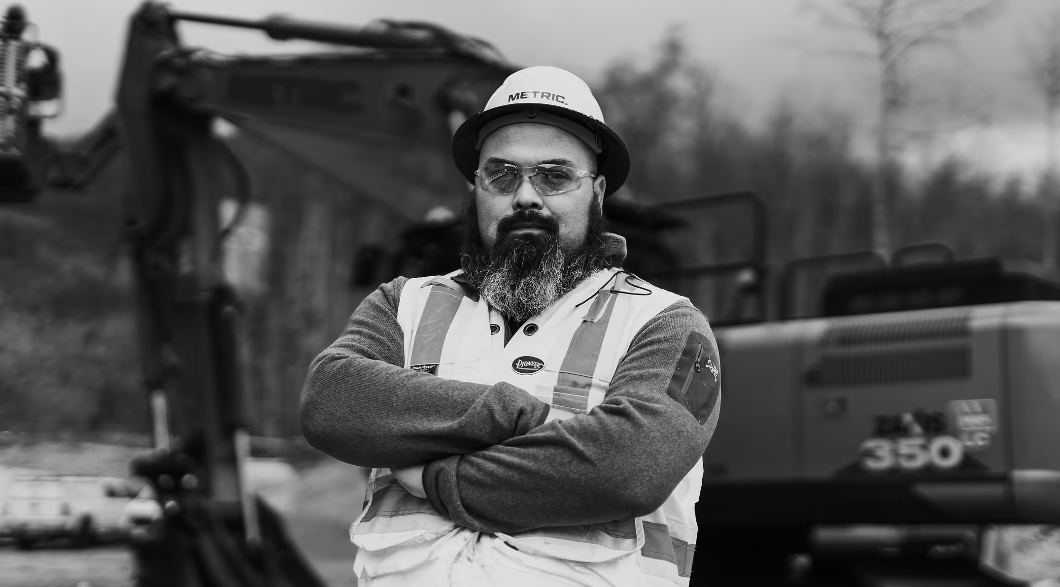 Confident construction worker with beard wearing hard hat and safety vest at a worksite with machinery.