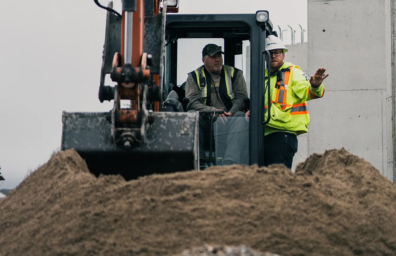 Two construction workers operating an excavator behind a large pile of dirt on site.