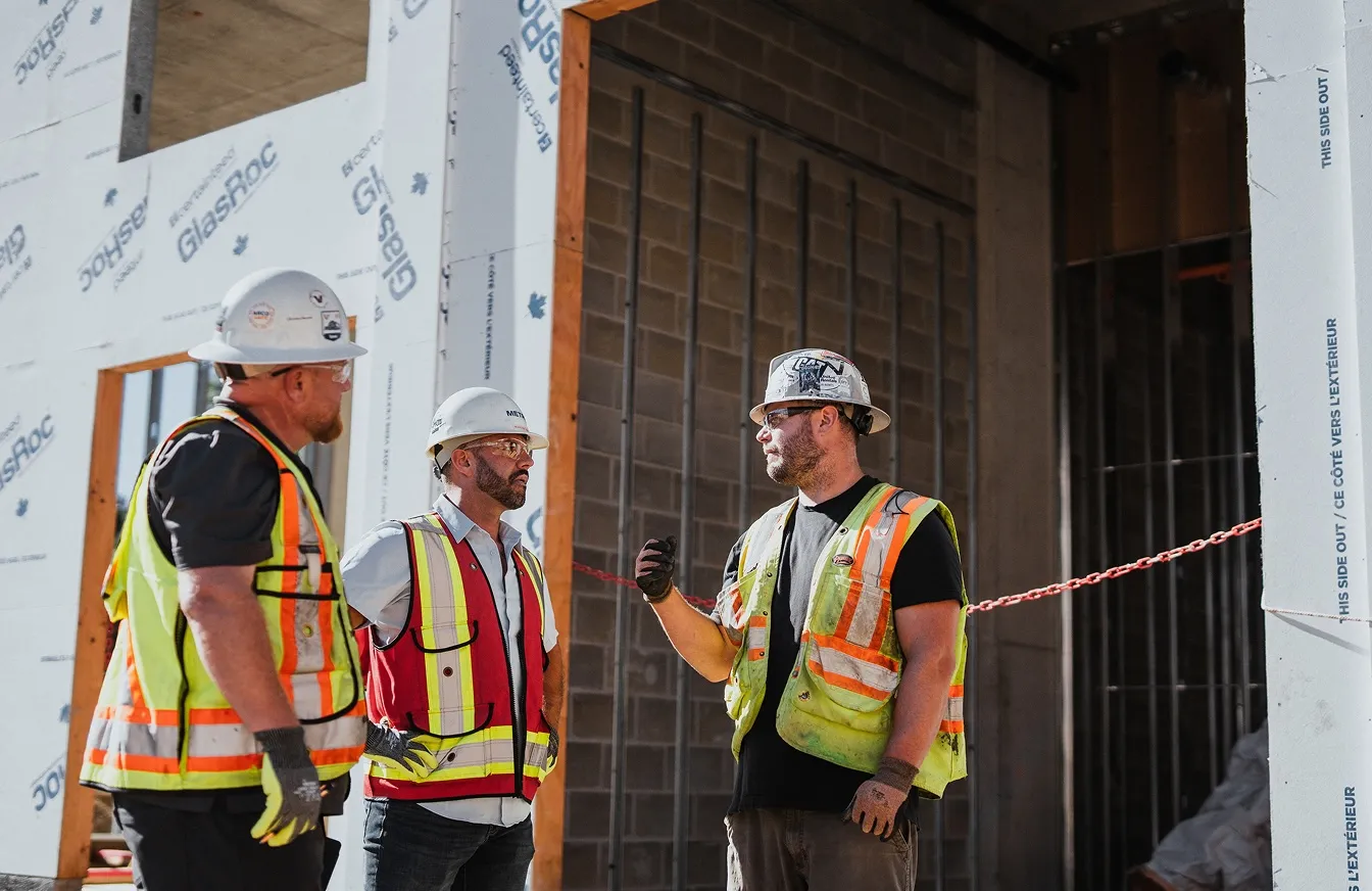 Three construction workers wearing hard hats and safety vests discuss work at a construction site.