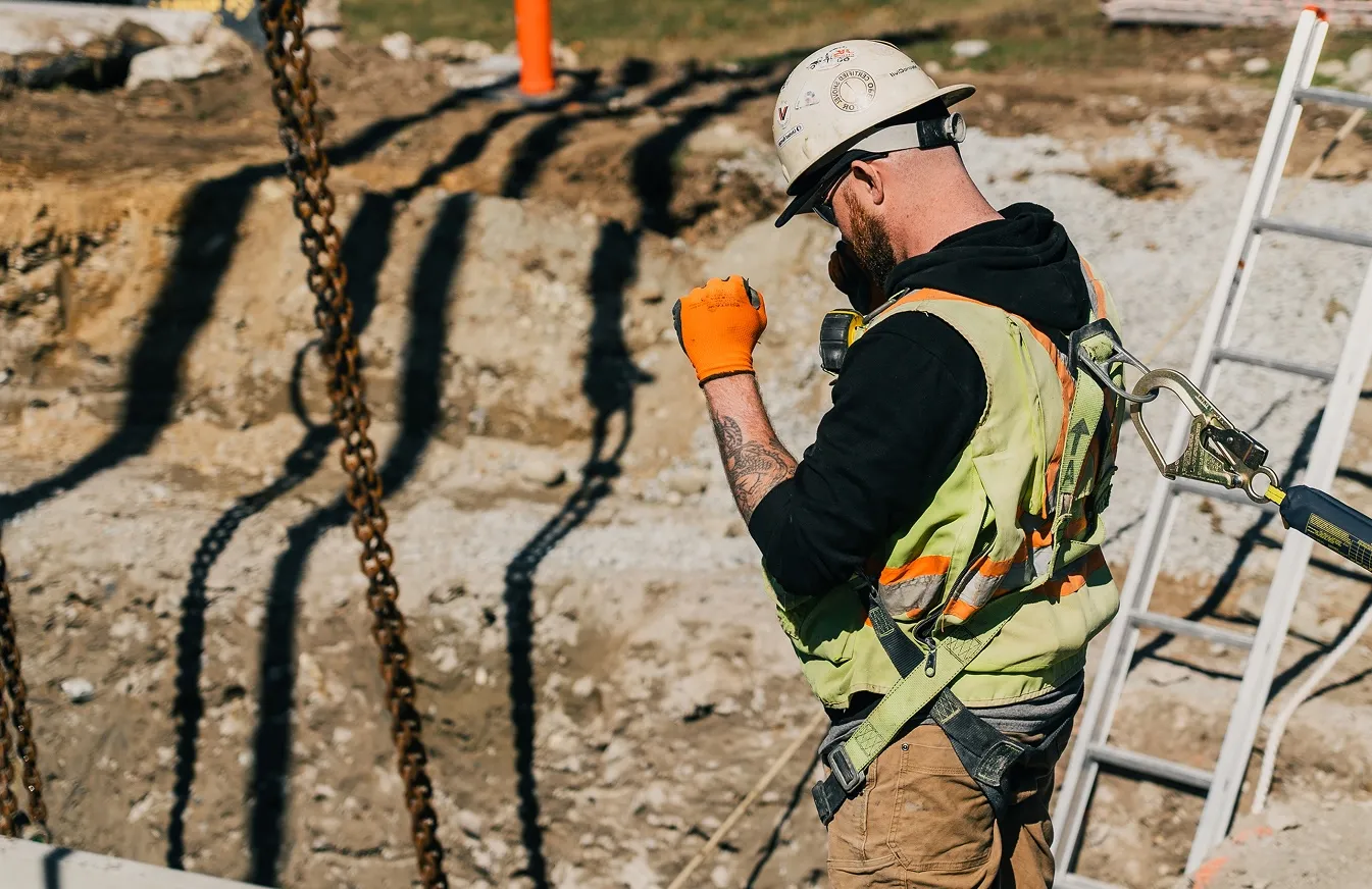 Construction worker with helmet, safety vest, and gloves on site using a phone and safety harness.