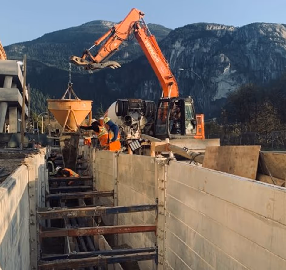 Construction workers operating an orange excavator and concrete mixer on a site with mountains in the background.