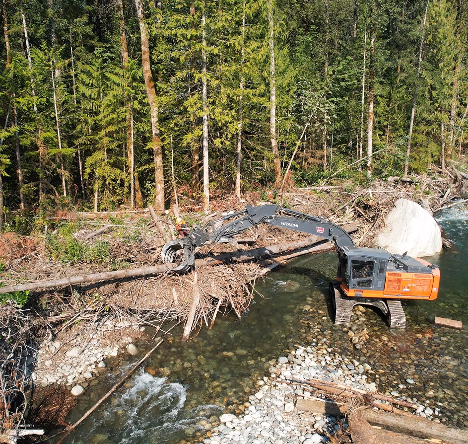 Excavator clearing logs and debris from a shallow forest river with rocky bed.