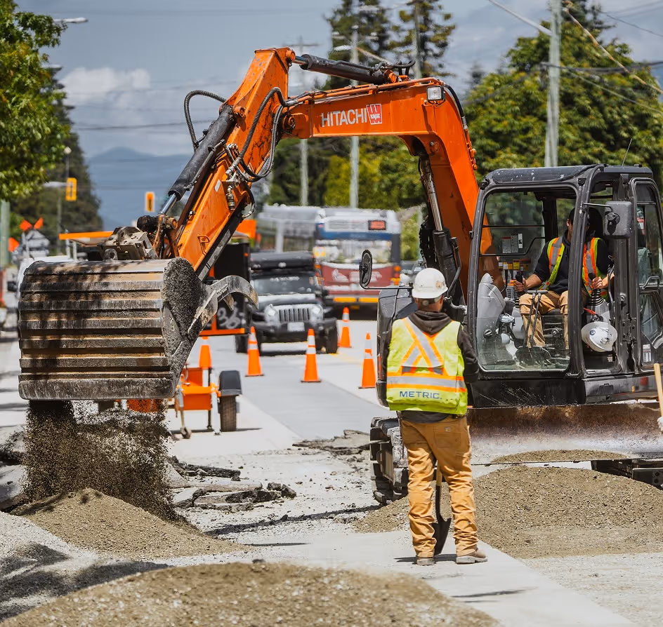 Construction workers operate an orange Hitachi excavator moving gravel on a roadwork site.