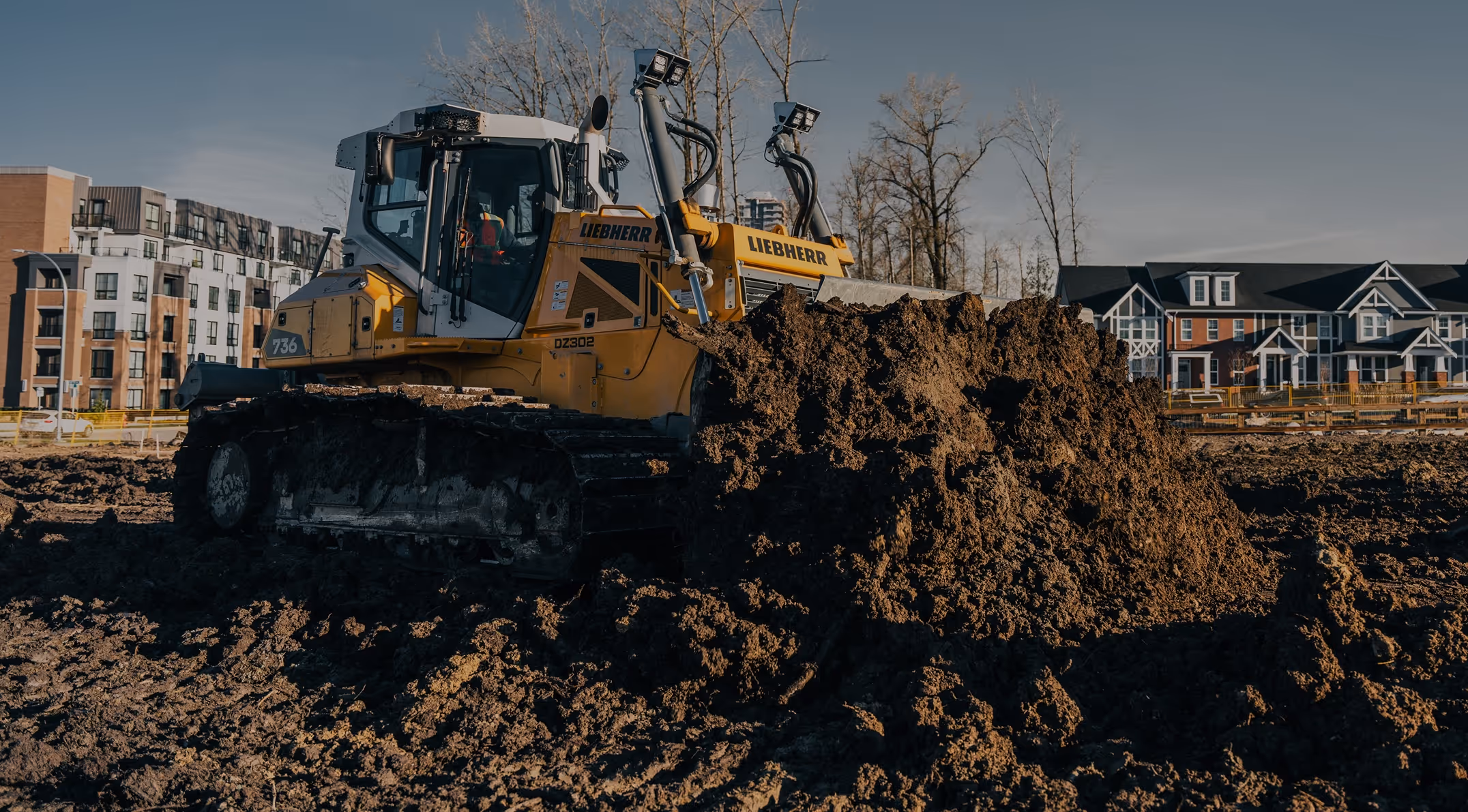 Yellow Liebherr bulldozer moving a large pile of dirt at a construction site near residential buildings.