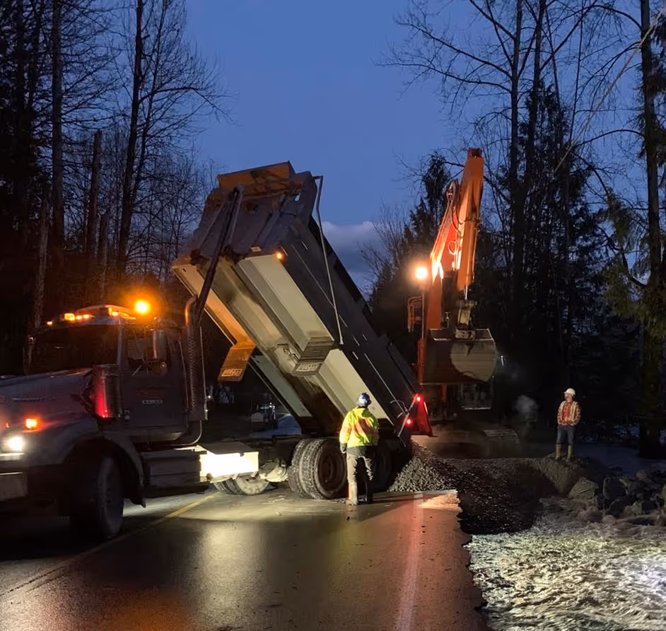 Construction workers and machines dumping gravel on a road at dusk near a forested area.