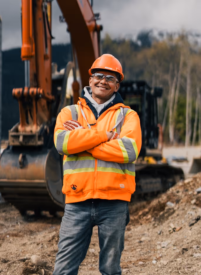 Smiling construction worker in orange jacket and helmet standing with arms crossed at a work site.