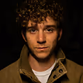 Portrait of a young man with curly hair wearing a brown jacket against a dark background.