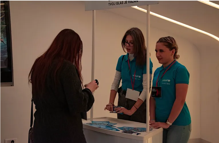 Dve devojke u plavim UNICEF majicama razgovaraju s posetiteljkom pored štanda.