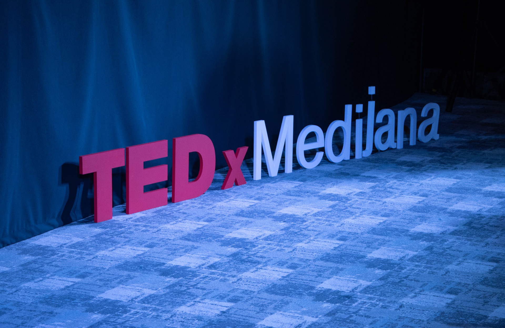 Veliki 3D natpis TEDx Medijana postavljen na plavi tepih ispred tamne pozadine.