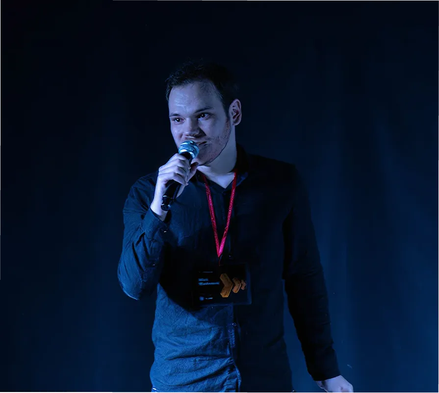 Mladi muškarac drži mikrofon i govori na sceni sa tamnom pozadinom.