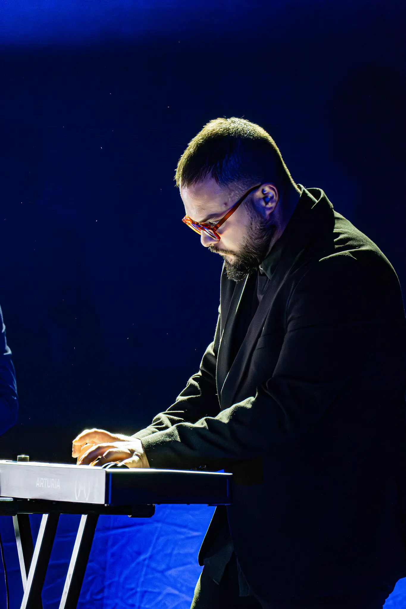 Muzičar sa kratkom kosom, bradom i naočarima svira elektronske klavijature marke Arturia na pozornici pod plavim svetlom. TEDx scena u Srbiji.