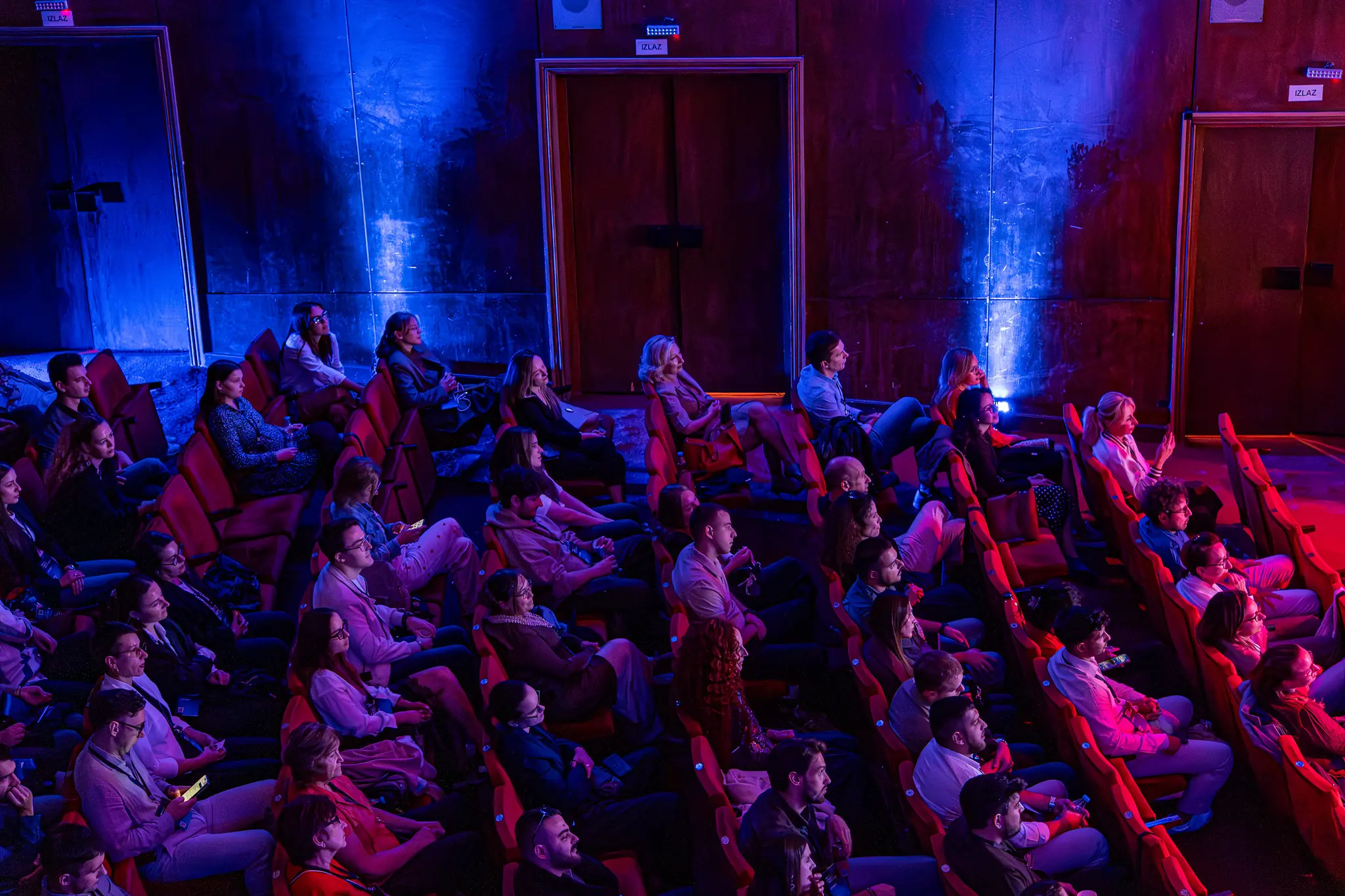 Ljudi sede u pozorišnim stolicama u sali sa plavim i crvenim osvetljenjem, fokusirani na događaj na bini ili ekranu. TEDx u Srbiji.