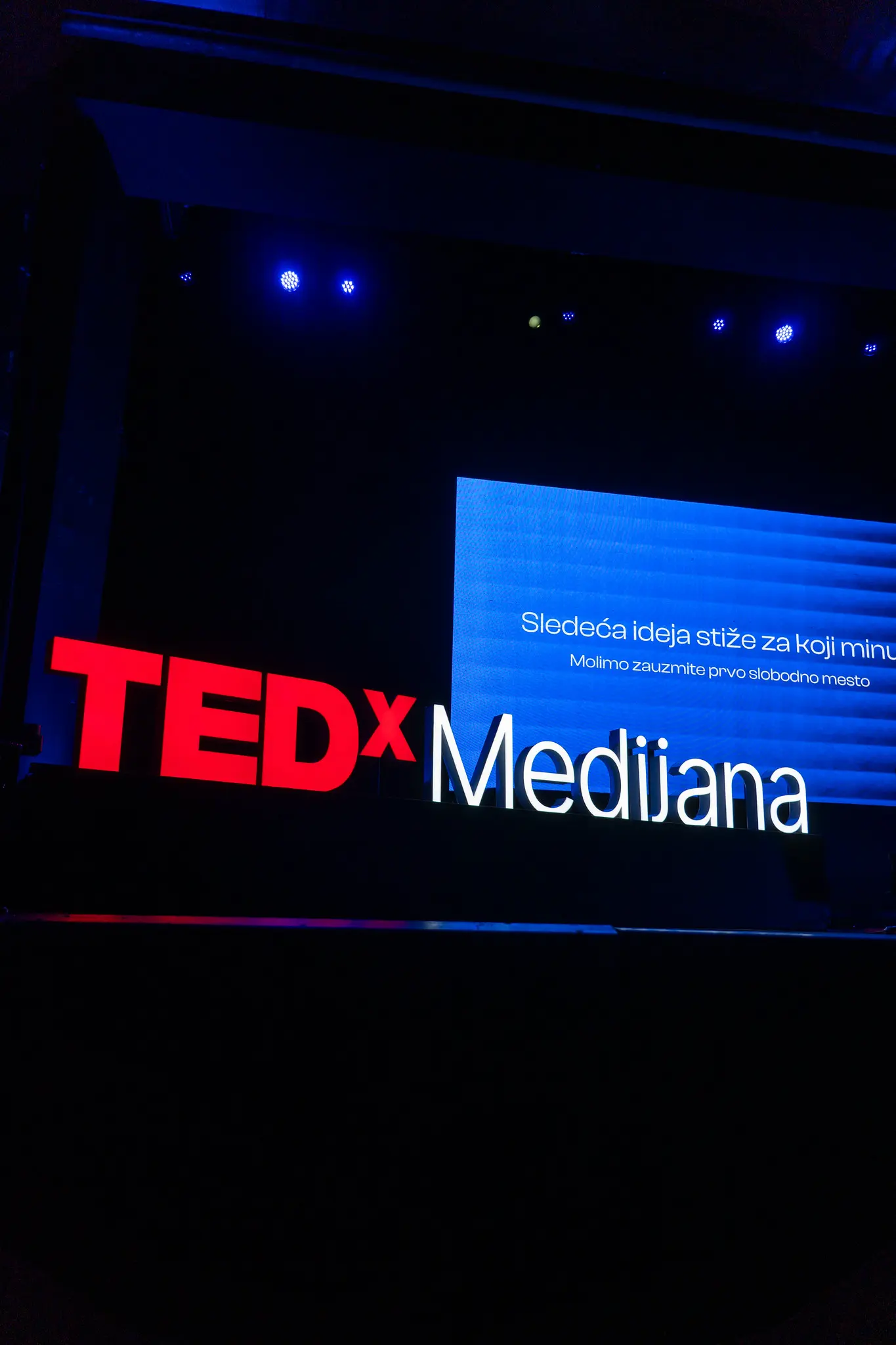 Osvetljeni natpis TEDx Medijana na sceni, sa plavim ekranom u pozadini na kojem piše na srpskom "Sledeća ideja stiže za koji minut. Molimo zauzmite prvo slobodno mesto." TEDx scena u Srbiji.