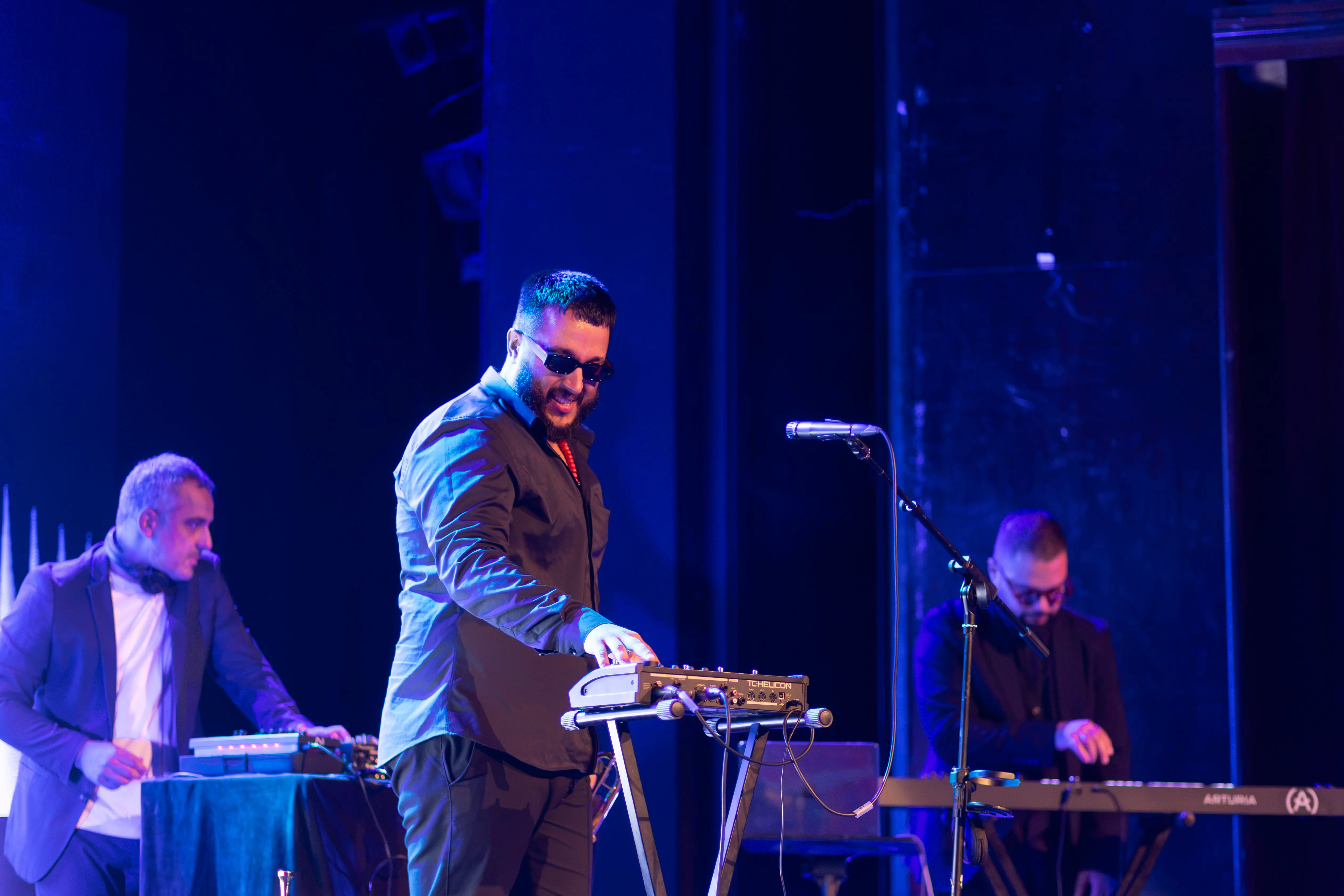 Tri muškarca na sceni tokom muzičkog nastupa, dva sviraju elektronske instrumente, jedan tastaturu, sa mikrofonom ispred, pod plavim svetlom pozorišta. TEDx scena u Srbiji.