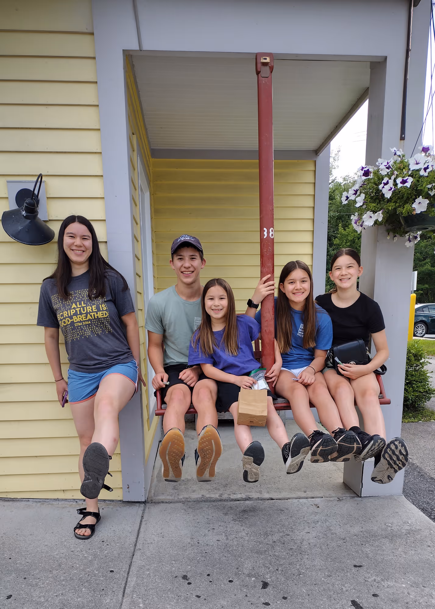 Five smiling young people casually sitting and standing on a red swing attached to a yellow building with white trim.
