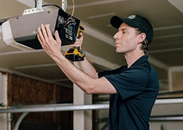 Technician installing or repairing a ceiling-mounted device indoors, wearing a black uniform and cap.
