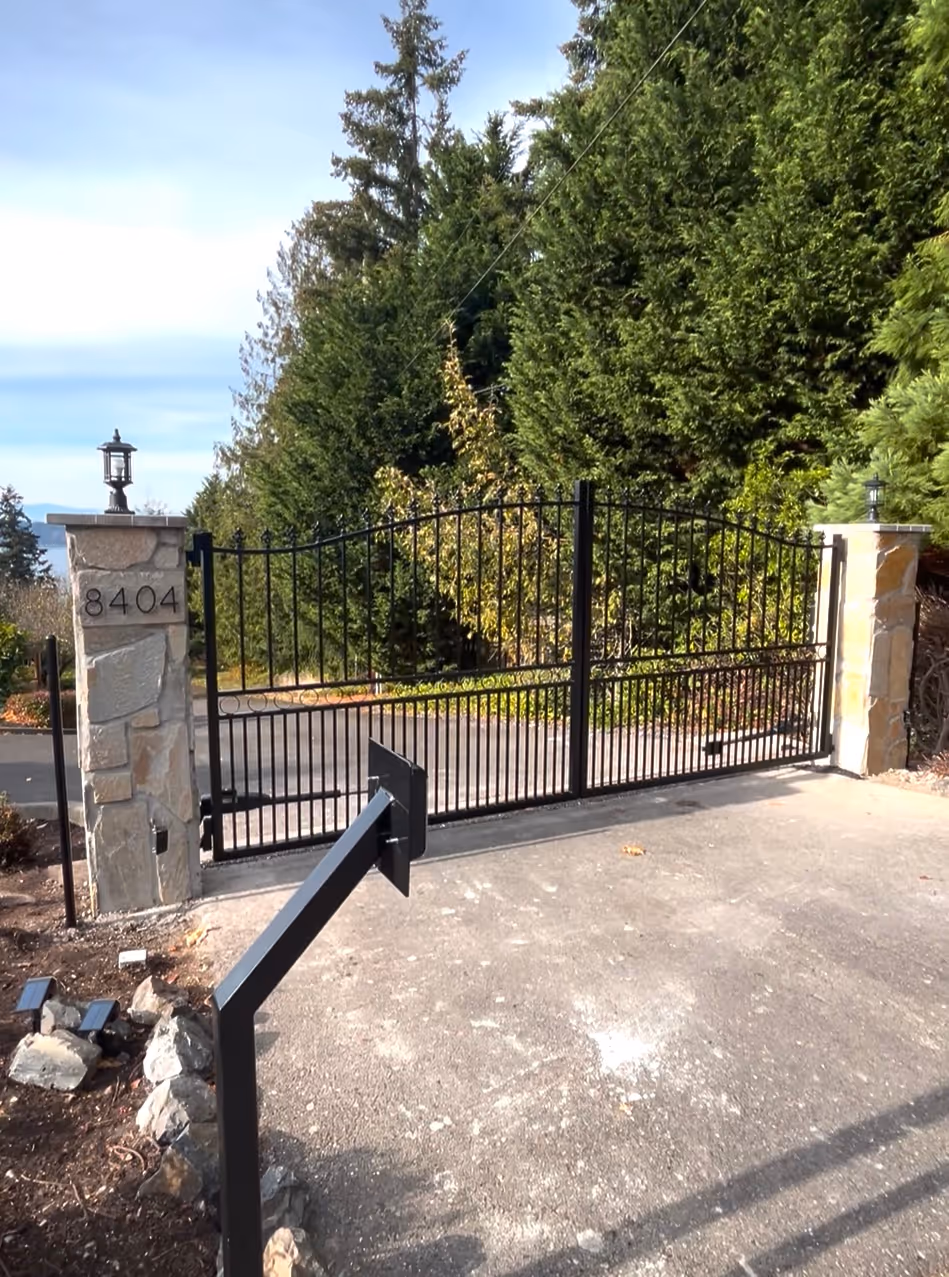 Black metal driveway gate between stone pillars with address 8404 and greenery behind.