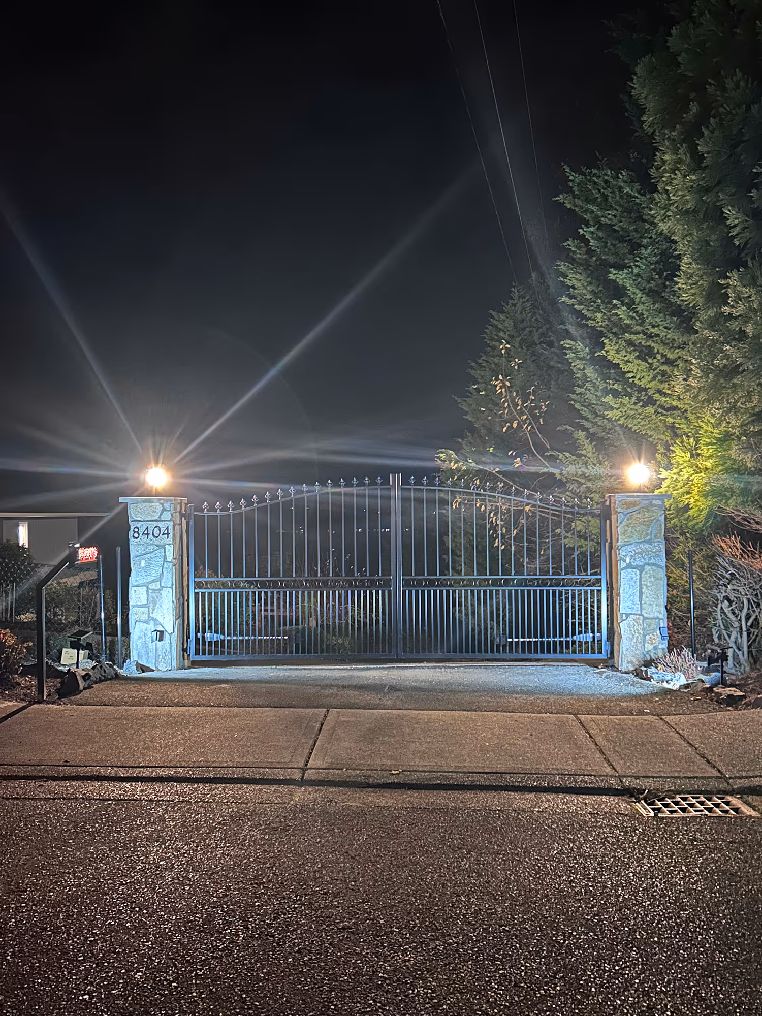 Closed metal gate at night with two stone pillars and bright lamps on each side, house number 8404.