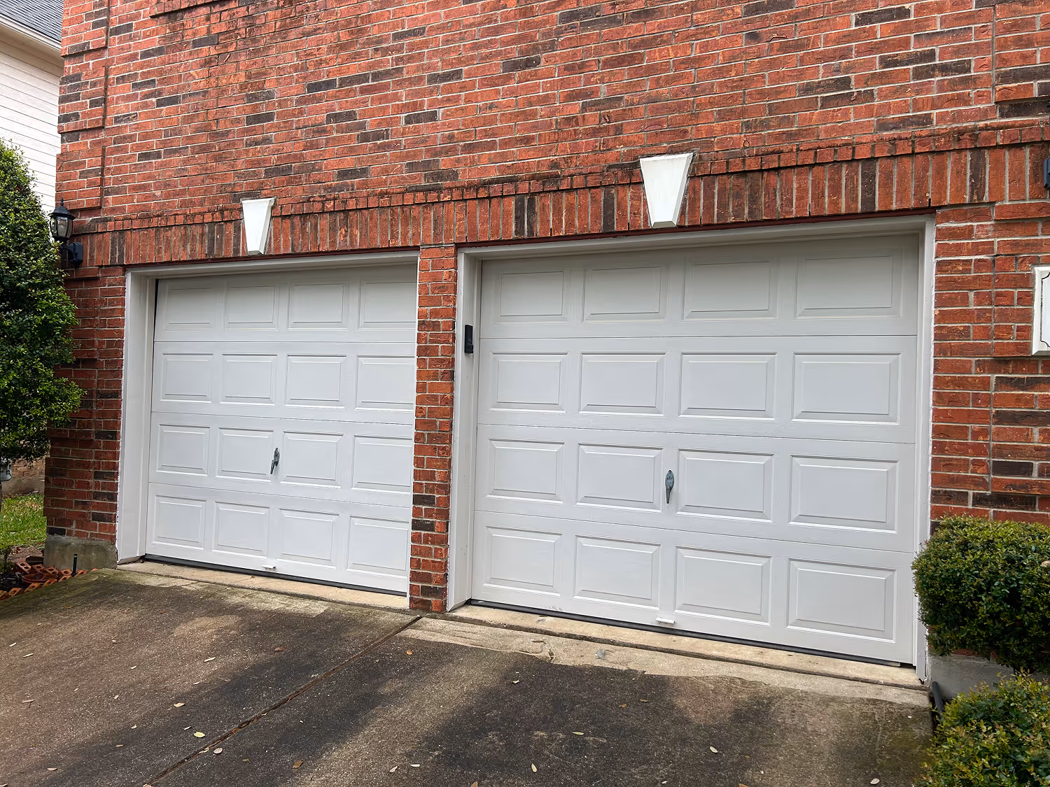 Two white garage doors with decorative panels set in a red brick house with concrete driveway.