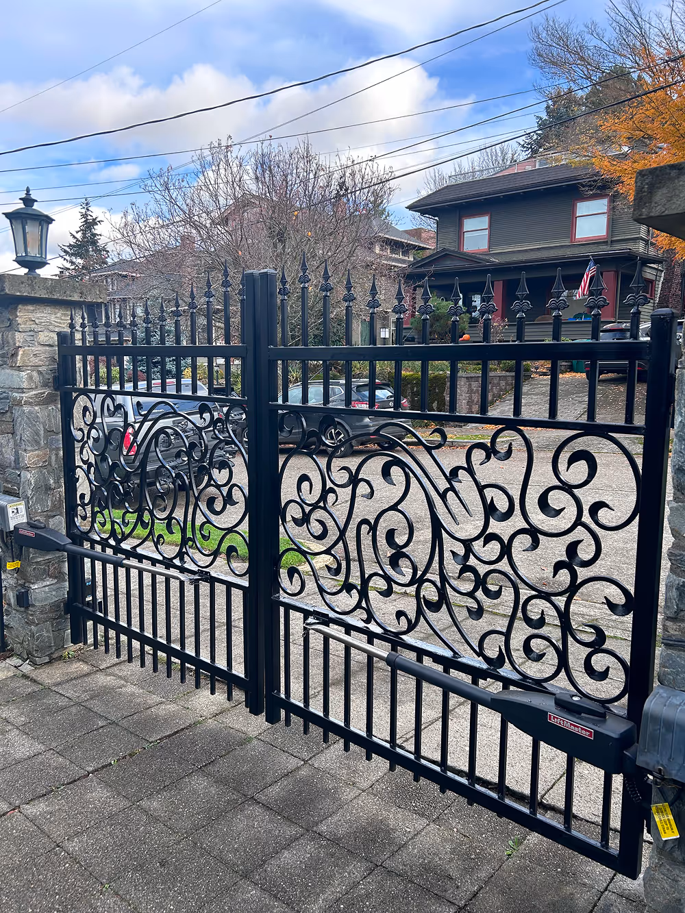 Black wrought iron gate with intricate scrollwork and automatic gate opener in front of a residential driveway and house.