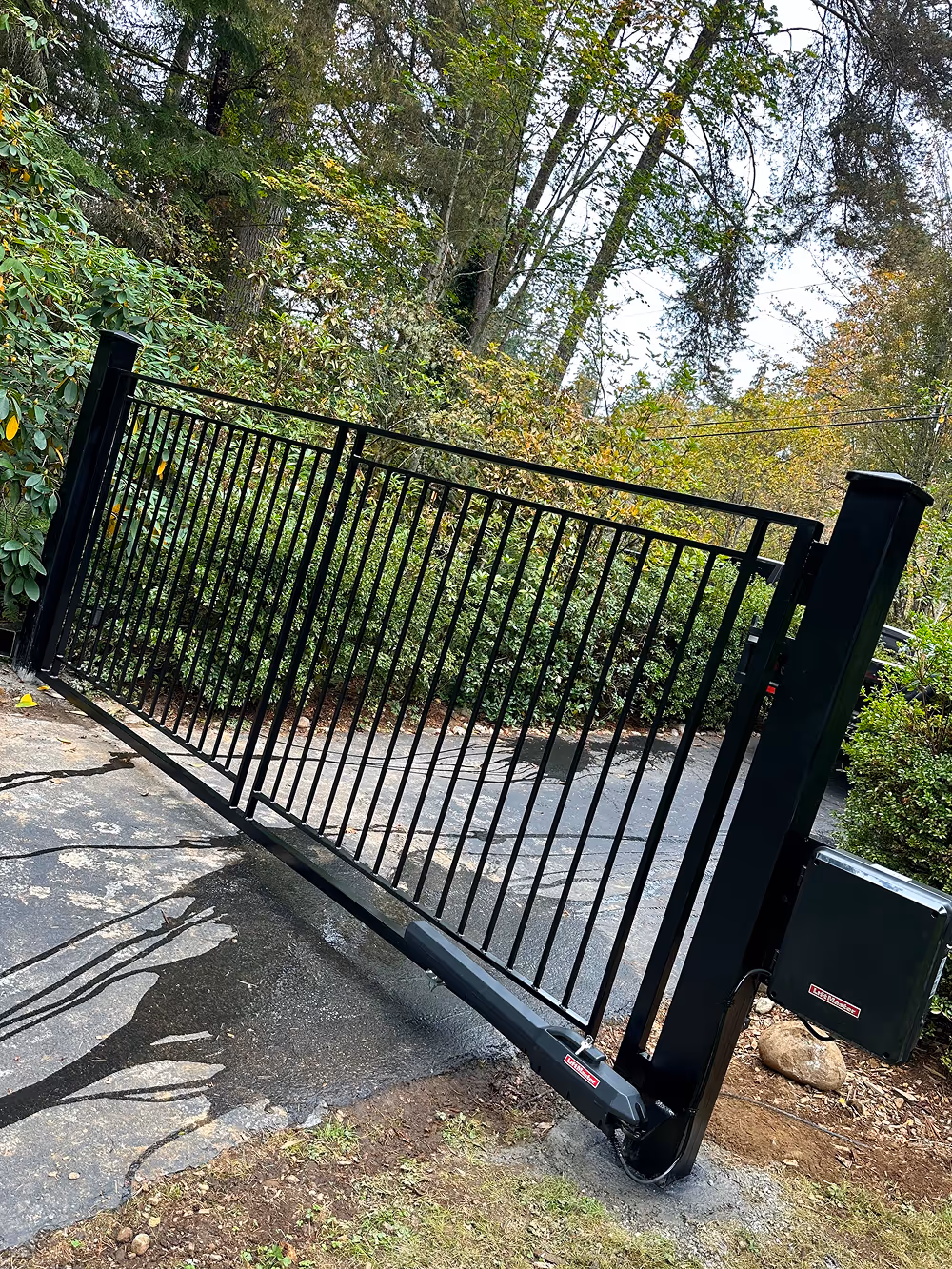Black metal automatic driveway gate partially open with surrounding green trees and bushes.