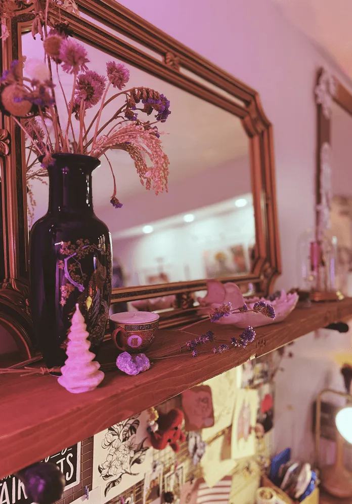 Wooden shelf with black vase holding dried flowers, a small ceramic cup, a white decorative item, and a mirror reflecting room details.