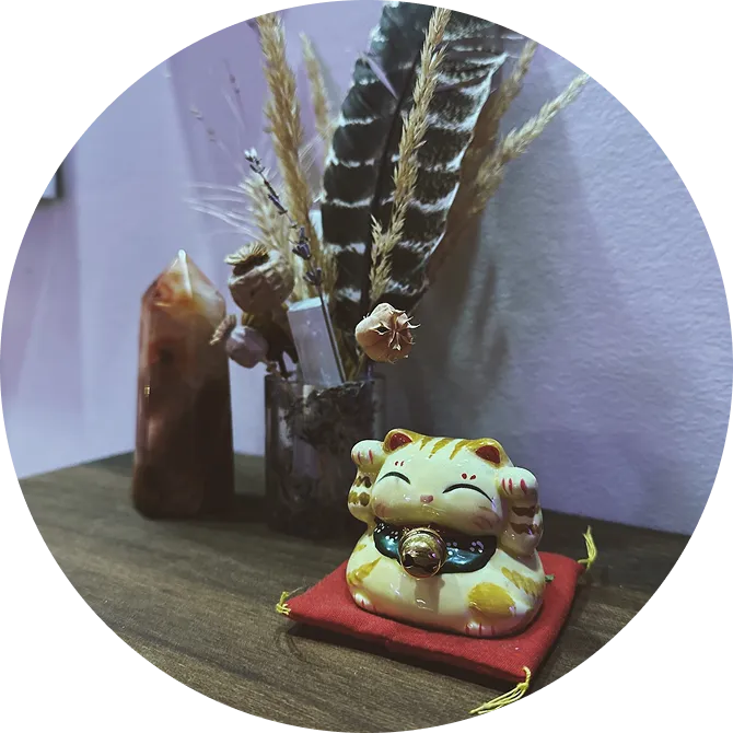Small yellow and white ceramic lucky cat figurine with a bell on a red cushion, sitting on a wooden surface next to dried plants and feathers in a glass container.