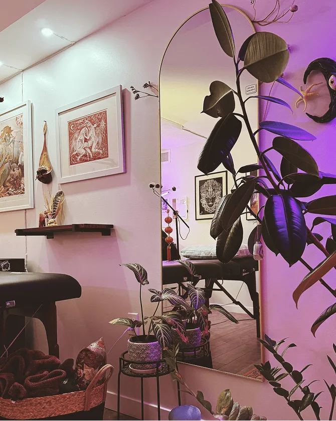 Cozy corner with a large arched mirror, potted plants on stands, framed artwork on the wall, and a massage table.